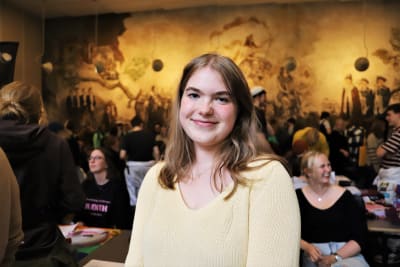 En ung kvinna med axellångt hår. I bakgrunden syns en vägg med målningar i gult och brunt föreställande studenter. 