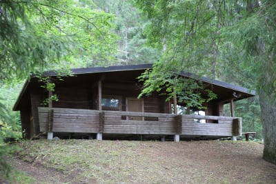 En mörkbrun bastubyggnad med veranda. Det går inte att se in i stugan eftersom gardinerna är fördragna. Bastun ligger mitt i skogen bland träd.