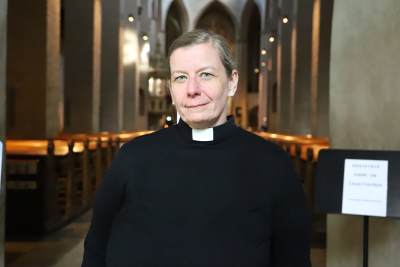 En kvinna med svart skjorta och prästkrage ser in i kameran.