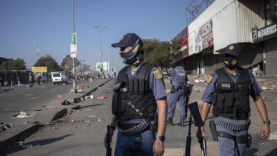 Polisen försöker stoppa plundring i förorten Alexandra i Johannesburg. Tre poliser står med hagelgevär utanför ett köpcenter med demonstranter i bakgrunden.