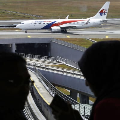 Varjossa olevat mies ja nainen keskustelevat, kun taustalla Malaysia Airlines -lentoyhtiön lentokone nousee kiitoradalla.