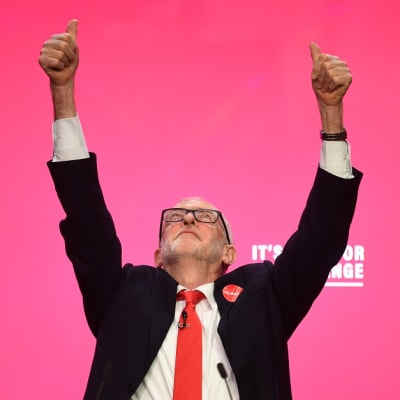 Kuvassa Jeremy Corbyn molemmat kädet ylhäällä ja katse ylöspäin suunnattuna. 