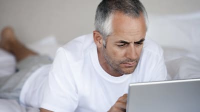 Medelålders man i vit t-skjorta ligger på en säng och ser på en dataskärm