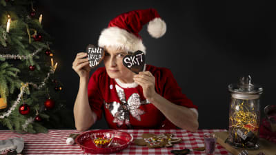 Annika Sylvin-Reuter sitter vid ett bord med glaserade pepparkakor. Hon är klädd i röd julklänning och tomteluva.  Hon håller upp två pepparkakor där det står "fans jävla jul" och "skit". . 
