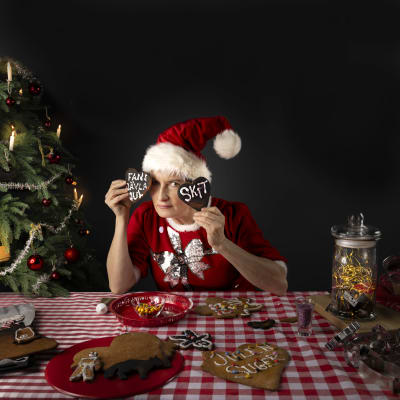 Annika Sylvin-Reuter sitter vid ett bord med glaserade pepparkakor. Hon är klädd i röd julklänning och tomteluva.  Hon håller upp två pepparkakor där det står "fans jävla jul" och "skit". . 