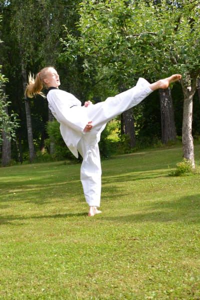 En ung kvinna i taekwondodräkt sparkar upp i luften. Hon står framför en röd husvägg.