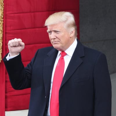 Trump seisoo punaisen vaatteen edessä oikea käsi kohotettuna, nyrkkiin puristettuna. Hänellä on tumma puku ja punainen kravatti.