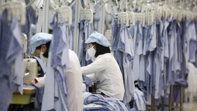 Bild på kvinnor som syr kläder i en klädfabrik. De bär munskydd och omges av blå tygbitar som hänger i ställningar ovanför dem.