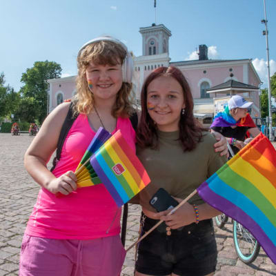 Två unga flickor står på Lovisa torg med regnbågsflaggor. De tittar in i kameran och ler.