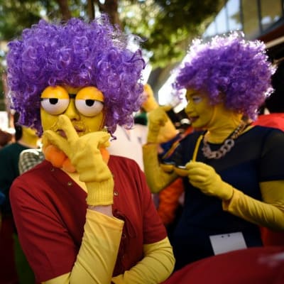 Kolmella halloweenin juhlijalla on keltaiseksi maalatut kasvot, violetit pörröperuukit ja tekomulkosilmät. 