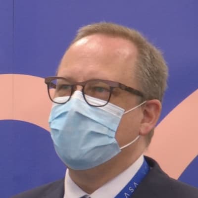 En man med munskydd och glasögon står framför en blå skärm.