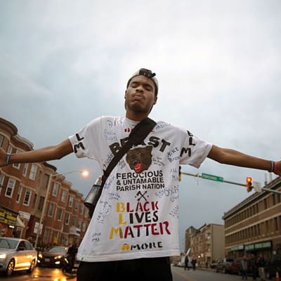 Nuori mustaihoinen mies on levittänyt kätensä ja seisoo keskellä katua. Hänellä on valkoinen t-paita, jossa on karhun pään kuva. T-paidassa lukee muun muassa "Black lives matter".
