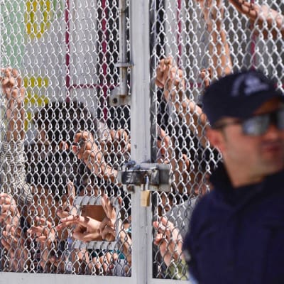 Turvapaikanhakijat ja siirtolaiset seisovat verkkoaidan takana. Oikeassa reunassa seisoo lippalakkipäinen univormupukuinen mies ja katselee oikealle.