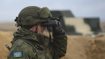 En soldat i kamouflagekläder tittar i en kikare.