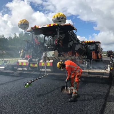 Tietyömaalla asfalttityöntekijä oranssissa haalarissa lapion kanssa taustallaan asfalttikone.