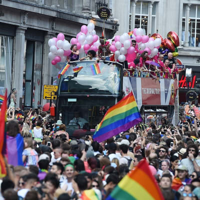 Katu on täynnä marssijoita, ja monet kantavat sateenkaaren värisiä lippuja. Kuvan keskiosassa keskellä ihmismerta on kaksikerroksinen bussi, jonka avoimessa yläosassa on ihmisiä valkoisten ja vaaleanpunaisten ilmapallojen kanssa.