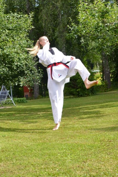 En ung kvinna i taekwondodräkt sparkar upp i luften. Hon står på en gräsmatta framför några äppelträd.