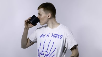 Edith Hammar står i en vit studio och dricker ur en kaffemugg, hon har en vit t-skjorta som det står "Du e homo" på.