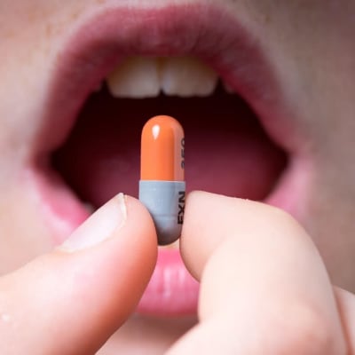 Lähikuva antibioottikapselista sormien välissä, taustalla ihmisen avoin suu. 