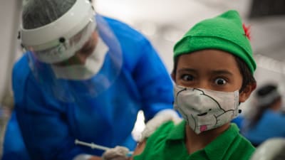 Ett barn i munskydd får coronavaccin av en sjukskötare i skyddsutrustning.