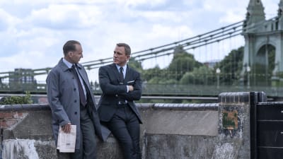 James Bond och M i samspråk stående vid en stenbro.