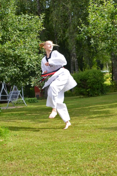 En ung kvinna i taekwondodräkt står ute på en gräsmatta. Hon håller på med att påbörja ett hopp.