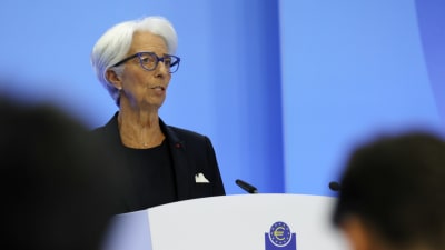 Christine Lagarde klädd i svart talar vid ett podium med texten European Central Bank - Eurosystem. I förgrunden syns huvuden av personer i publiken.
