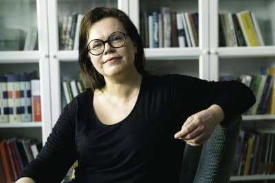 På bilden en brunhårig kvinna med glasögon som sitter framför en inglasad bokhylla. 