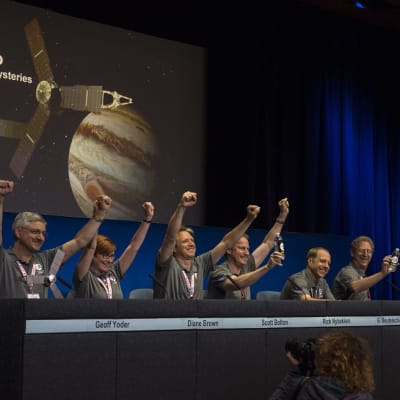 Joukko Juno-työryhmän jäseniä pitää käsiään voitonriemuisesti ylhäällä tiedotustilaisuudessa pöydän takana. Kaikilla on harmaat T-paidat. Taustalla on kuvaesitys luotaimesta ja planeetasta.