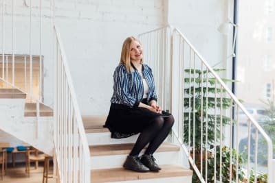 Michaela von Kugelgen sitter i en trappa. Väggen bakom är vit och trappan är också vit, med trappräcke av metall. Hon har på sig en svart kjol och en blåspräcklig jacka. 