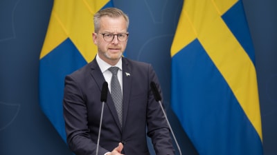 Inrikesminister Mikael Damberg vid regeringskansliets presspodium med svenska flaggor i bakgrunden.
