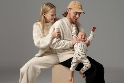 Muotoilijapari Saana Sipilä ja Olli Sallinen istuvat selät vastakkain vanerikuution päällä. Olli pitelee pariskunnan vuoden ikäistä vauvaa sylissään. Studiokuva.
