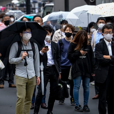 Ihmisiä katukäytävällä rinta rinnan. Kaikilla on maskit. Osa kantaa sateenvarjoa. 