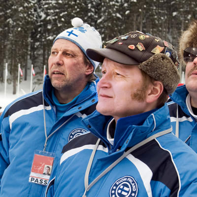 Suomen hiihtomaajoukkueen asuissa olevat miehet katsovat intensiivisesti urheilusuoritusta.