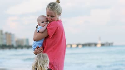En kvinna håller om ett spädbarn på en sandstrand medan hon ser mot en annan liten flicka som går bredvid.