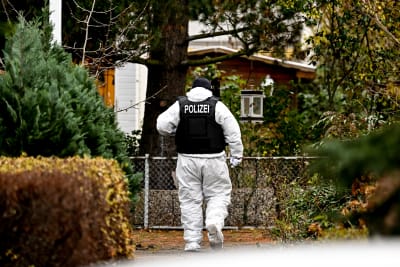 En polis i vit skyddsdräkt och mörk skyddsväst med texten Polizei på ryggen går mellan häckar i ett småhusområde i höstfärger.