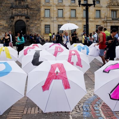 Joukko ihmisiä maalaa värikkäitä kirjaimia valkoisiin sateenvarjoihin, jotka on asteltu torille. Edesä olevien kolmen sateenvarjon kirjaimista muodostuu sana "paz", rauha. 