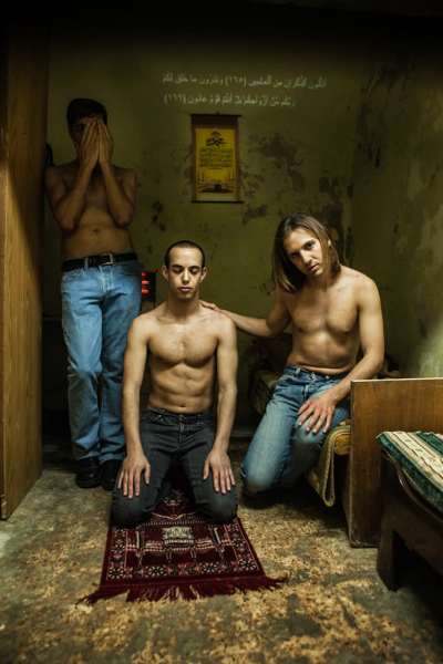 Tre män med bara överkroppar i ett litet rum, två står och den tredje knäböjer på en bönematta.
