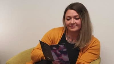 Kvinna med blont hår, tatueringar och stickad gul tröja sitter och läser en broschyr.
