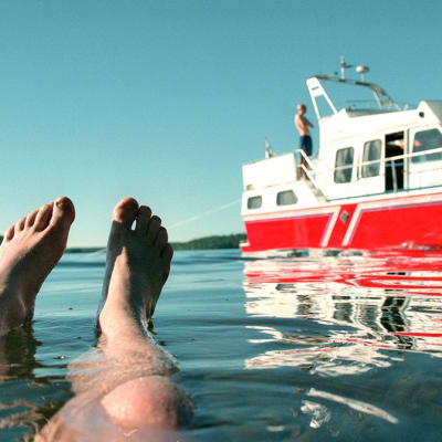 Miehen jalat veden pinnalla, taustalla punainen vene rantautuneena.