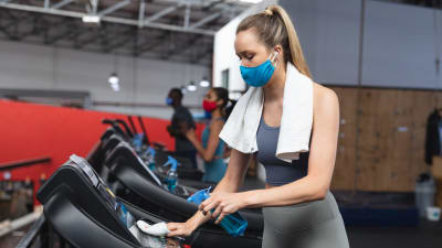 En kvinna i munskydd torkar av en löpmatta med en handduk på gymmet.