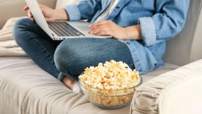 En person sitter på en soffa med dator i famnen. Bredvid henne syns en skål med popcorn.