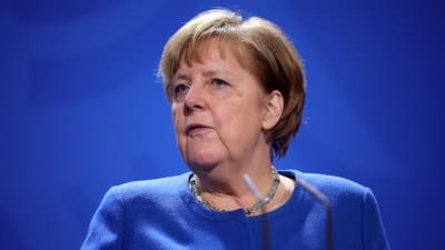 Angela Merkel, blå tröja framför en blå bakgrund. Framför mikrofoner.