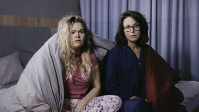 Två kvinnor sitter i en säng, med pyjamas på, och ser uppgivna ut. 