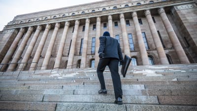 En person i svarta kläder och väska går upp för trapporna till riksdagshuset i Helsingfors.