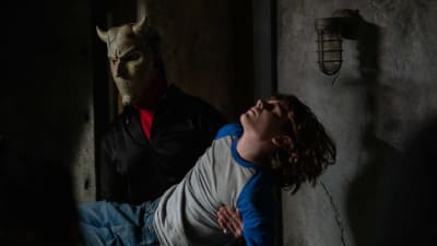 En maskerad figur bär en medvetslös pojke i ett skumt rum. 
