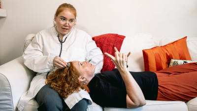Heidi ligger i soffan med huvudet i Vivis knä. Vivi håller ett medicinskt stetoskop mot Heidis tinning som under en undersökning.