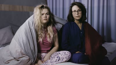 Två kvinnor sitter på en säng iklädda pyjamas och ser förskräckta ut. Ett täcke hänger över ena axeln på kvinnan till vänster.