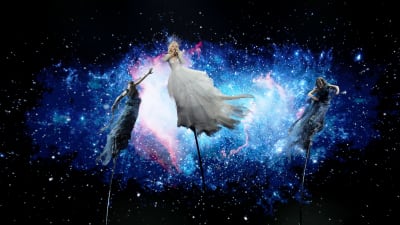 kate som sjunger för australien står tillsammans med två dansare iklädd i svart och själv i vit klänning på tre cirkuspålar med en bild av universum i bakgrunden