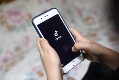 En person håller i en vit telefon. I skärmen syns Tiktoks logo; svart bakgrund med en not och texten "Tiktok".